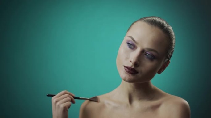 化妆申请的步骤。蓝眼睛的女孩使用风扇制造的化妆刷在她的脸和身体上涂上荧光笔。