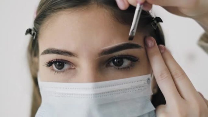 大师在美容院用医用口罩给一名年轻女子画眉毛的形状。特写