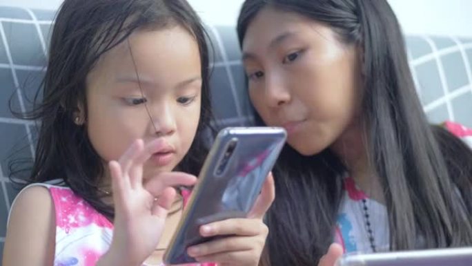 快乐的亚洲儿童使用智能手机和数字平板电脑观看病毒视频和玩网络游戏，同时在家一起在沙发上放松，生活方式