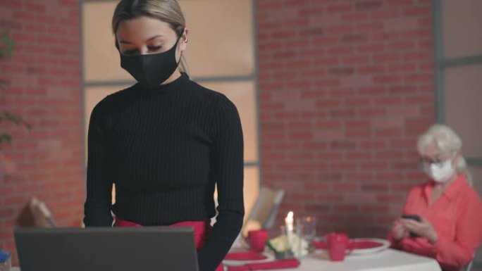 一名顾客坐在后台看着她的手机，而一名戴着口罩的年轻女服务员进入餐厅的POS计算机系统进行销售。