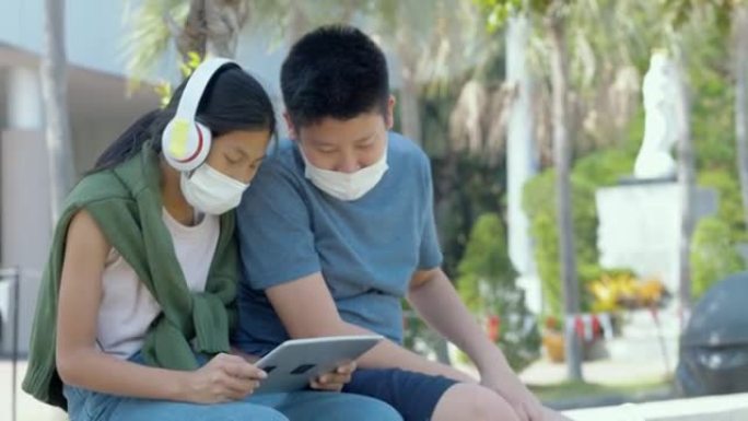 快乐的亚洲学童戴着口罩，在校园户外用数字平板电脑和耳机放松，社交疏远的生活方式理念。