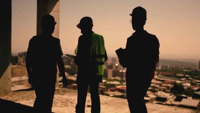 一群戴着防护头盔的专业工程师在未完成的建筑中行走和交谈。城市的后视图。穿着防护服的男人在高层多层建筑