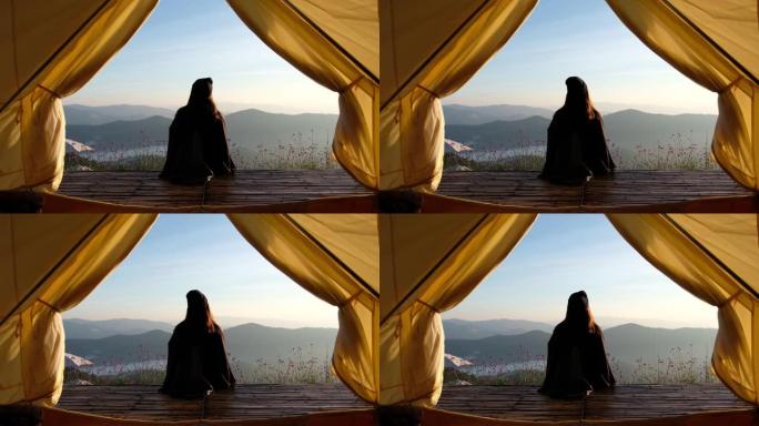 一个女人坐在木制阳台上看日出和帐篷外美丽的山景的后视