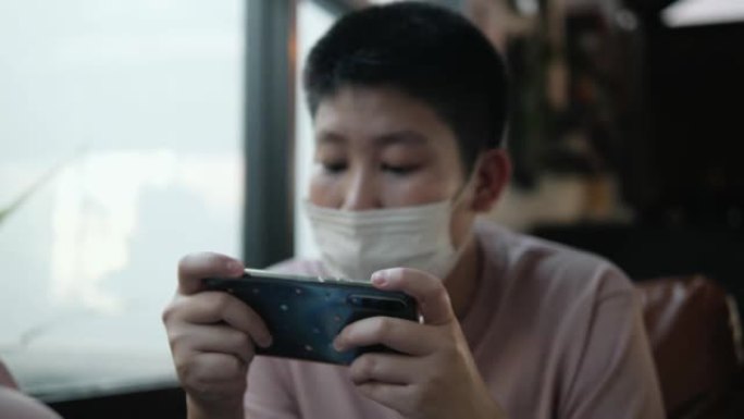 强调亚洲男孩戴着口罩，在餐厅窗口附近用智能手机玩网络游戏，生活方式概念。