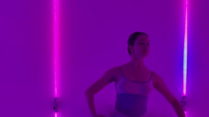 女人在霓虹灯的背景下在工作室里跳芭蕾舞并表演舞蹈元素。女孩以垂直的裂口抬起腿，然后进行转弯。慢动作。