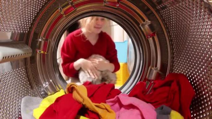 有趣的猫坐在洗衣机里，从洗衣机里看。女孩从洗衣机里带走猫