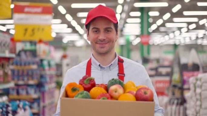 快乐的年轻送货员或穿着红色制服的快递员拿着纸板箱，里面放着新鲜蔬菜，站在超市里。快速快递安全送餐网购