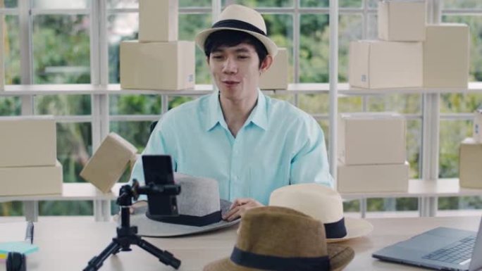微笑的亚洲男性影响者回顾软呢帽，并在新型冠状病毒肺炎大流行期间在家在线销售产品。