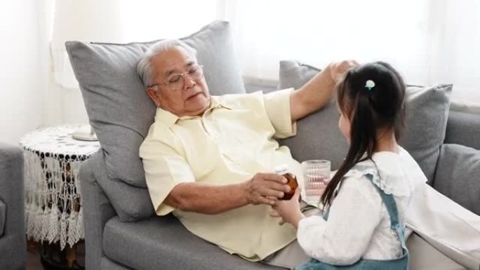幸福的家庭小亚洲女儿带着药物和一杯水给生病的爷爷在家里的沙发上照顾，老人病人退休享受生活理念。
