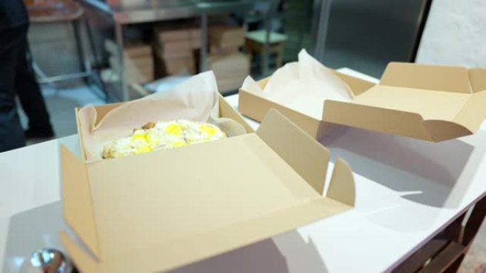 男厨师将煮熟的烤糕点放在一次性纸盒中交付。无法识别的高加索人在餐厅或比萨店厨房准备外卖的美味食物