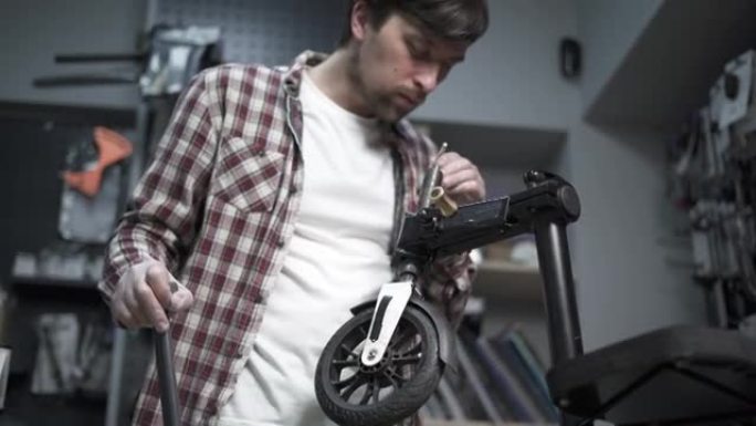 年轻的白人男性机械师在修理厂工作，修理孩子们踢踏板车。修理滑板车，维修服务。生态运输。在特殊车间修理