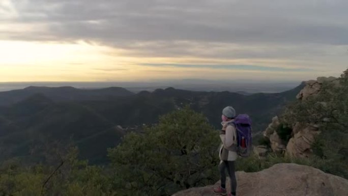 登山妇女在山顶上戴着帽子，背包和大流行面具看着地平线