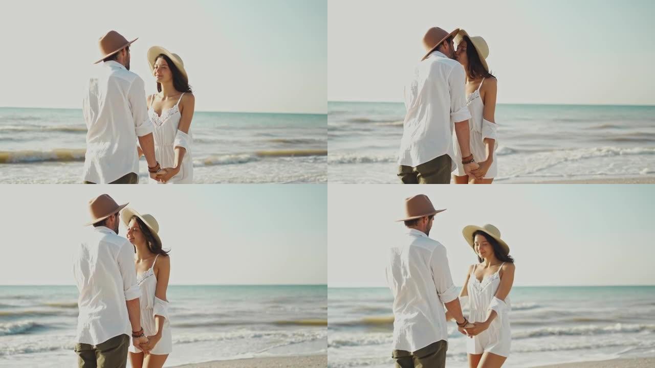 充满爱心的年轻夫妇戴着帽子凝视着彼此的眼睛，在阳光明媚的早晨手拉手一起站在沙滩上时亲吻