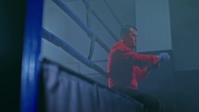 年轻的男拳击手正坐在烟雾笼罩的黑暗大厅里的拳台上，为训练做准备。拳击手用保护性绷带包住他的手和手指。