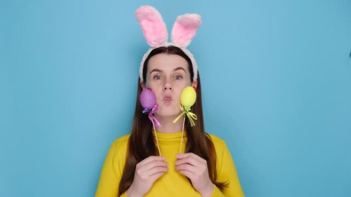 可爱的漫画年轻女子戴着兔子耳朵，做鬼脸，玩傻瓜，拿着彩色鸡蛋，表情愉快，穿着毛衣，蓝色背景上的模特，