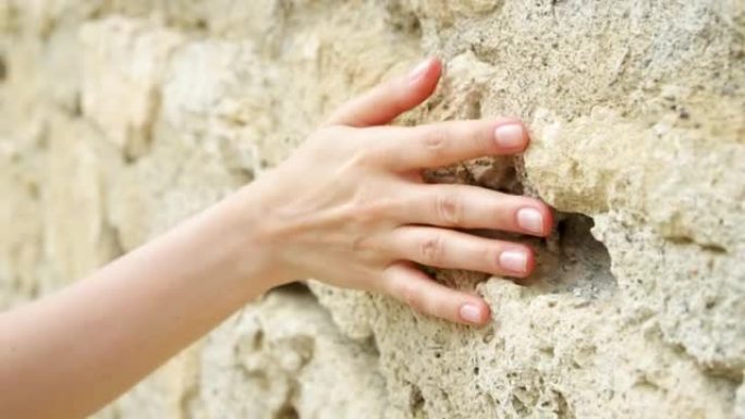 女人在慢动作中滑手靠在旧石墙上。女性手触摸岩石粗糙表面