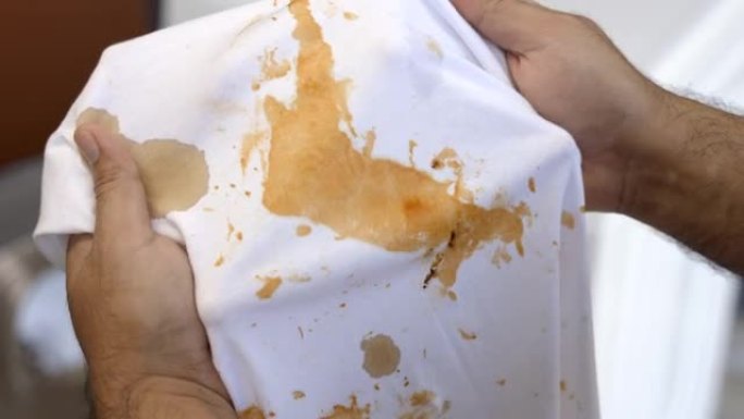 白色棉衬衫有很多污渍和污垢。沾满番茄酱的男人的手握着衬衫，打开它看污垢。