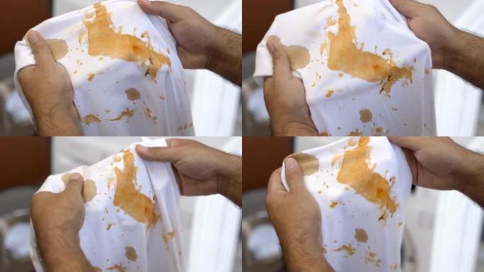 白色棉衬衫有很多污渍和污垢。沾满番茄酱的男人的手握着衬衫，打开它看污垢。