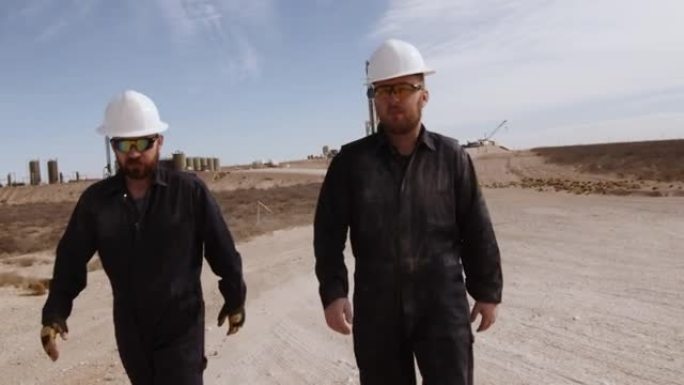 两名年轻的油田工人穿着PPE阻燃工作服和安全帽，从西德克萨斯州的压裂油气钻机走开