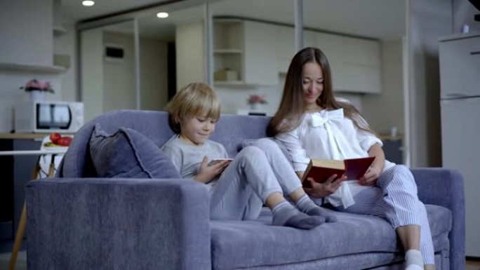 放松身心的小男孩在平板电脑上看卡通，因为模糊的微笑的女人坐在背景沙发上看书。快乐无忧无虑的儿子和母亲
