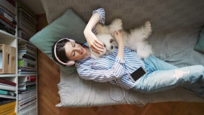 年轻女子躺在沙发上，一边听音乐一边和她的狗玩耍的俯视图