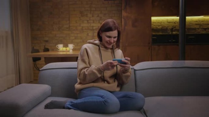 年轻的成年女性在手机上玩手机游戏，坐在沙发上赢得游戏。女性在智能手机上玩时用手做胜利手势。