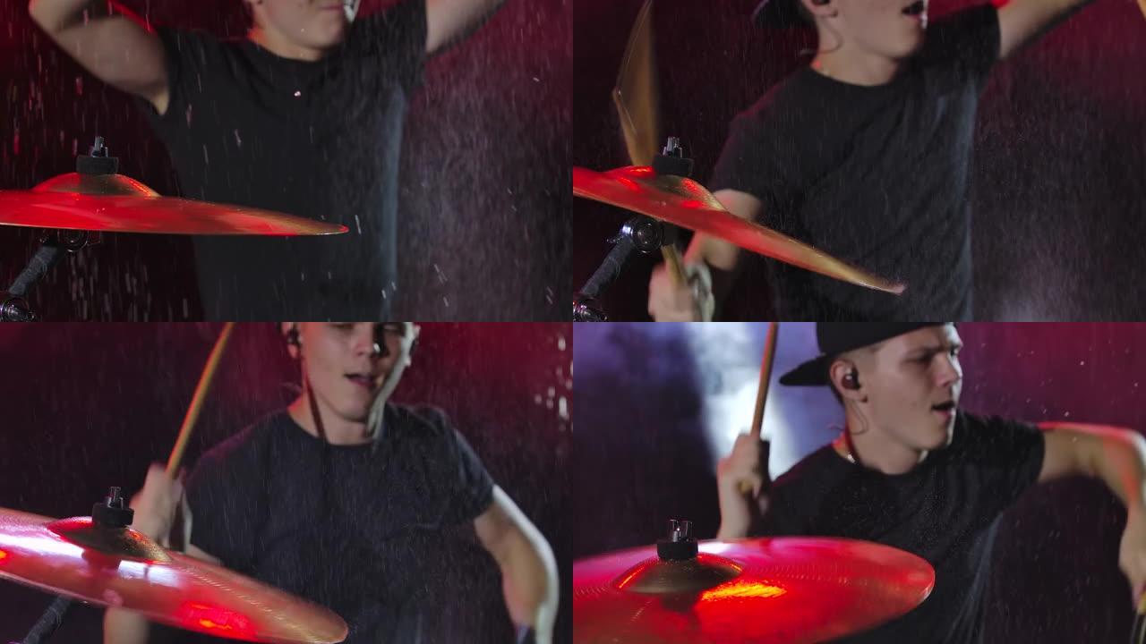 年轻的摇滚音乐家在雨滴中的黑暗工作室演奏鼓乐。人击打钹和鼓，吐了很多喷雾。红光着色的模糊背景。特写。