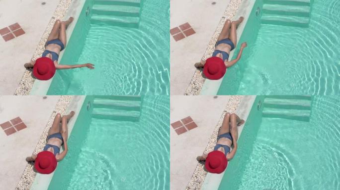 年轻的美女躺在水疗度假酒店的游泳池旁放松。炎热的夏日和明亮的阳光。享受晒黑和放松与大红帽子盖面部保护
