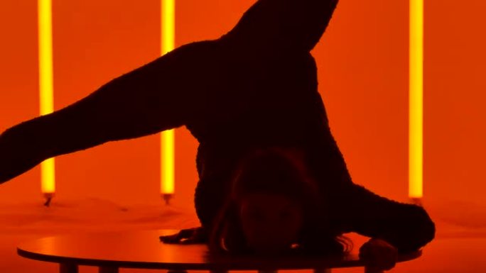 一个有魅力的女人表演倒立，移动她的腿，把它们分成一个裂口。工作室里的体操运动员在五彩霓虹灯的背景下。