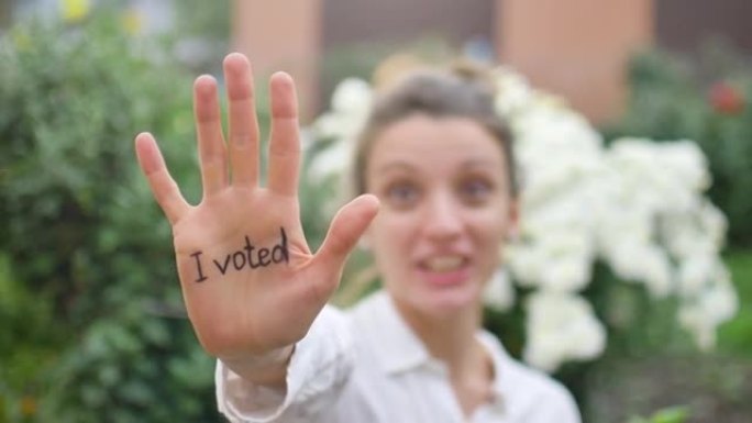 户外女性肖像的年轻成年妇女在白色衬衫显示她的手与口号“我投票了”的鲜花背景，选举日的概念