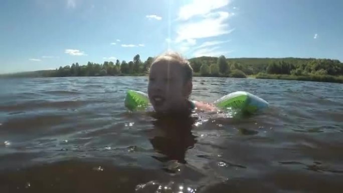 一个无所畏惧的小女孩的肖像，她在明亮的太阳的背景下，在远离海岸的河上游泳，穿着充气臂章。特写。从水面