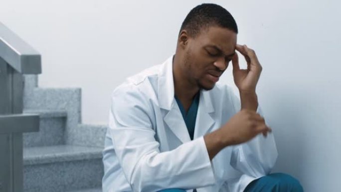 医护人员在新型冠状病毒肺炎期间的压力。疲倦的黑人医生坐在医院的楼梯上，头痛