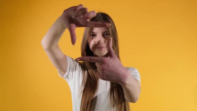 女孩用手指框住她的脸，假装拍照，橙色的工作室背景。特写