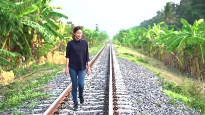 年轻漂亮的亚洲女人穿着黑色衬衫和吉恩把头发绑在后面，在当地的铁路上快乐地笑着，看着边路的香蕉树森林