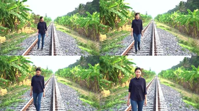 年轻漂亮的亚洲女人穿着黑色衬衫和吉恩把头发绑在后面，在当地的铁路上快乐地笑着，看着边路的香蕉树森林