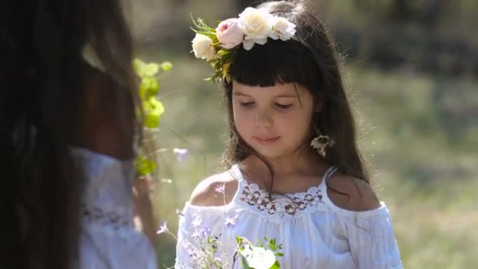 小高加索学龄前女孩的特写肖像，长长的棕色头发和蓝色的眼睛在花环上看着她的姐姐在自然背景上收集花束。4