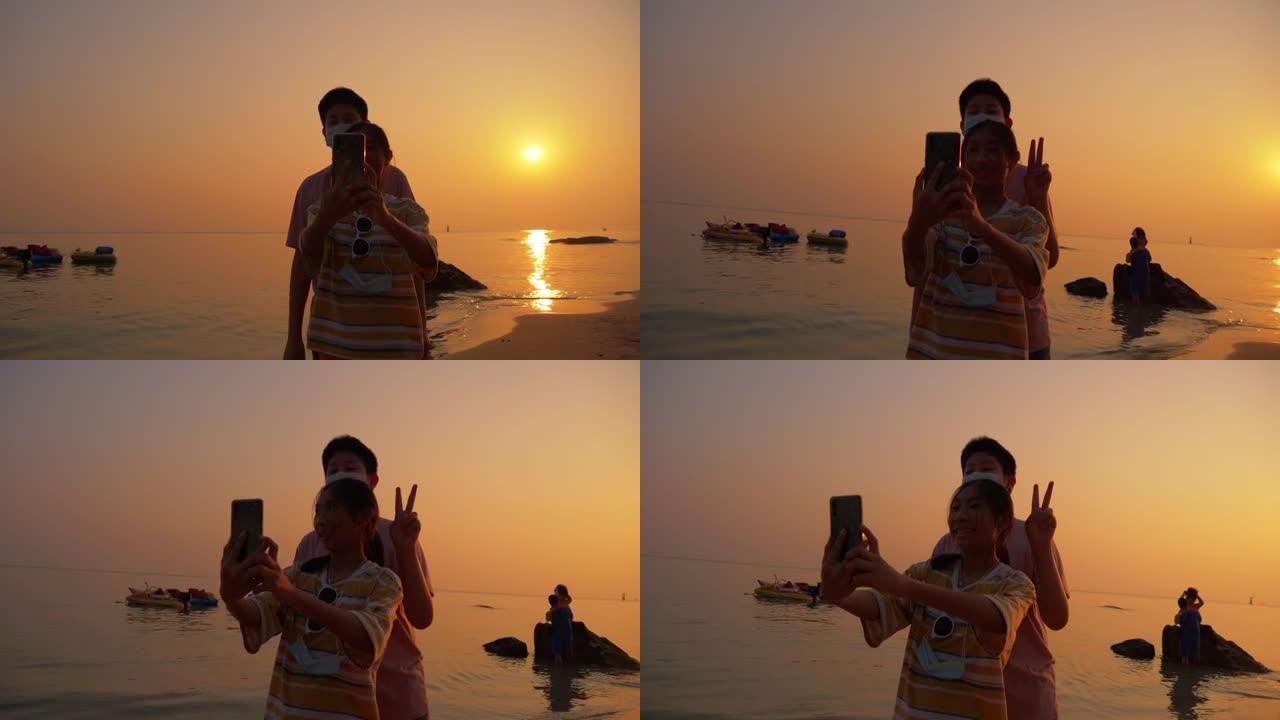 儿童在海滩上拍照和自拍的剪影与日出背景，生活方式概念。