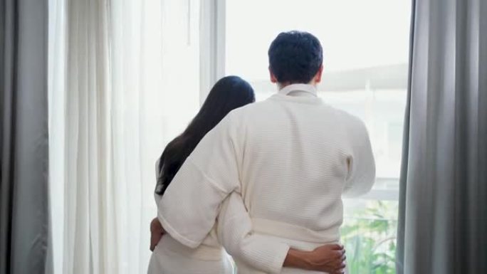 穿着浴袍的亚洲夫妇早上在豪华酒店卧室醒来时打开窗户上的窗帘