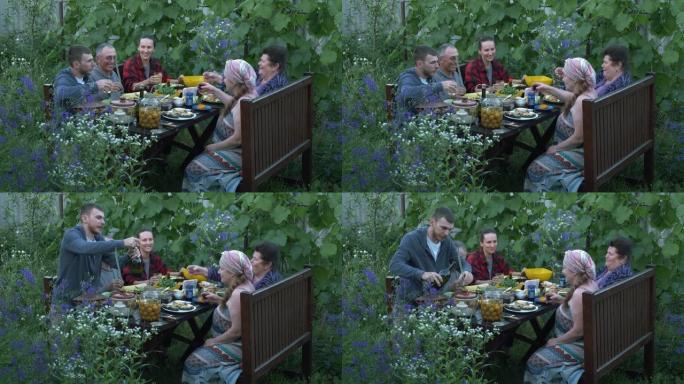 在户外野餐时聚集亲戚和朋友。年轻和年迈的亲戚在院子里的餐桌上吃喝。家庭一起吃饭，玩得开心。幸福的家庭