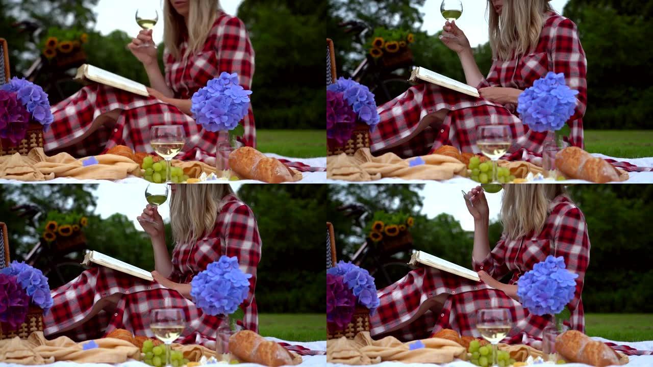 穿着红色方格连衣裙和帽子的女孩坐在白色针织野餐毯上看书，喝酒。阳光明媚的夏天野餐，有面包、水果、花束