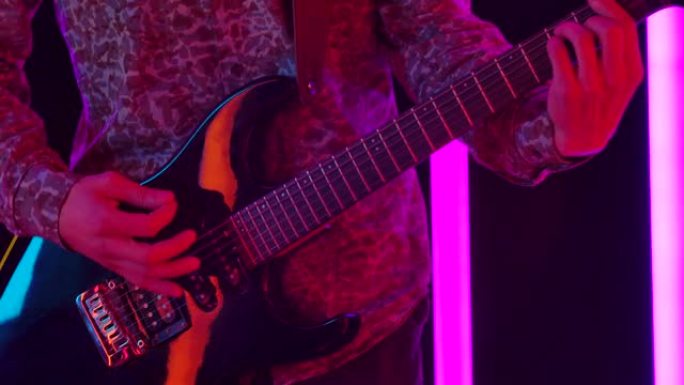 在明亮的彩色霓虹灯管的背景下，吉他手在工作室中充满活力的表演。一个男人的双手紧紧拉紧黑色电吉他的琴弦