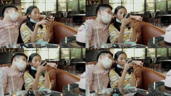戴着口罩的亚洲男孩看着他的妹妹在智能手机上观看病毒视频，同时在餐厅放松，生活方式概念。