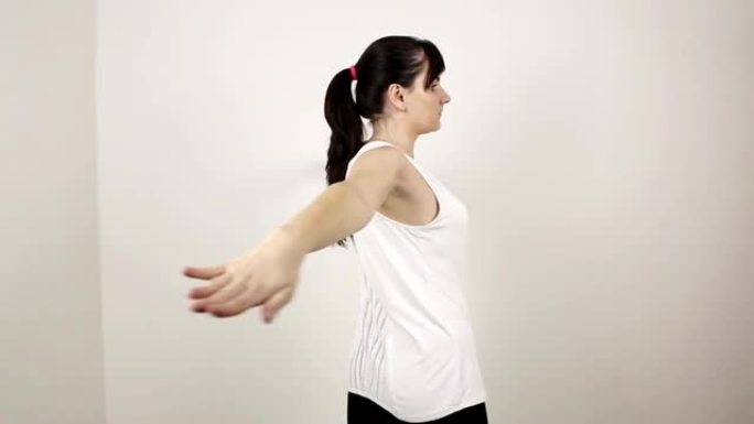 年轻漂亮的黑发白人女孩穿着白色背心在家做体操。为胸部进行手臂摆动运动，健康