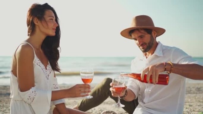 穿着白色衣服的夫妇在户外约会，喝酒野餐，坐在沙滩上的毯子上，可以看到海景。男人在杯子里倒粉红色的酒，