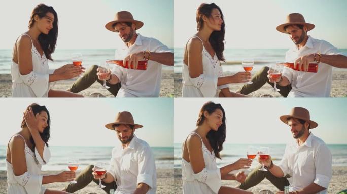 穿着白色衣服的夫妇在户外约会，喝酒野餐，坐在沙滩上的毯子上，可以看到海景。男人在杯子里倒粉红色的酒，