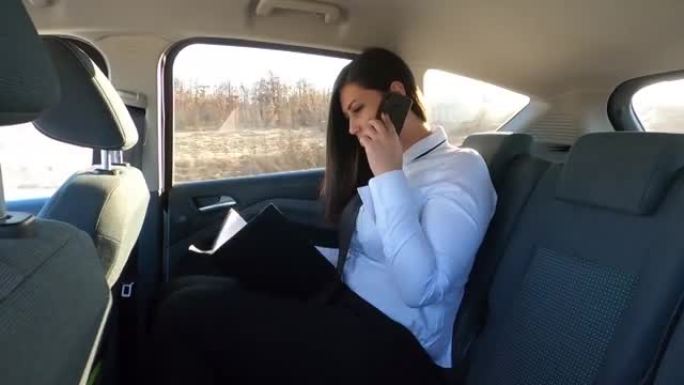 一位年轻的女商人坐在汽车的后座上-她正在出差的路上，他使用平板电脑电话，翻阅文件，进行约会并在旅行时