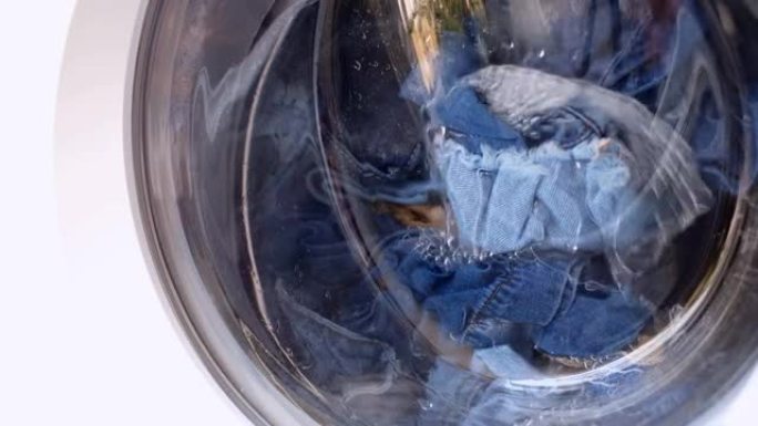 衣物在玻璃门后面的白色自动洗衣机中洗涤，金属滚筒依次沿不同方向旋转。洗衣服牛仔裤。家庭作业4k