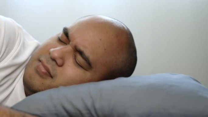 穿着白色t恤的30岁左右的亚洲秃头男人睡不好觉。他有头痛、失眠和噩梦。
