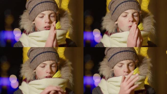 在城市里，被冻结的高加索男孩暖手摩擦手掌和吹户外的特写镜头。除夕背景上带有金色圣诞灯的可爱孩子的肖像
