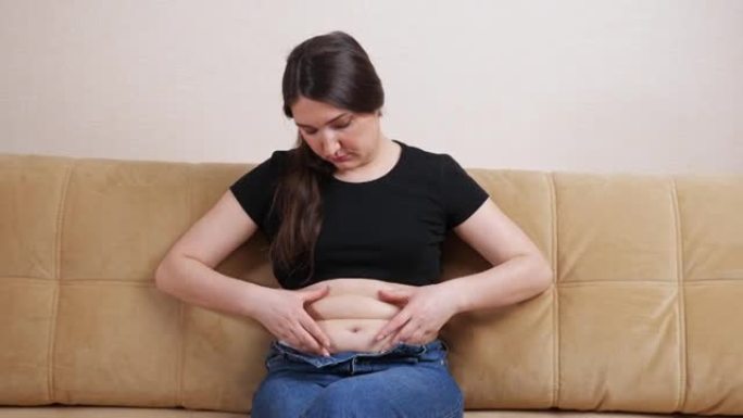 年轻女子坐在沙发上时感到肚子上的脂肪褶皱